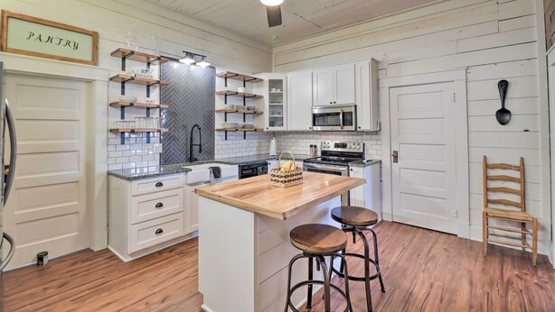 Una cocina con armarios blancos, una isla de madera con taburetes, un ventilador de techo y un cartel que dice 'PANTRY'.