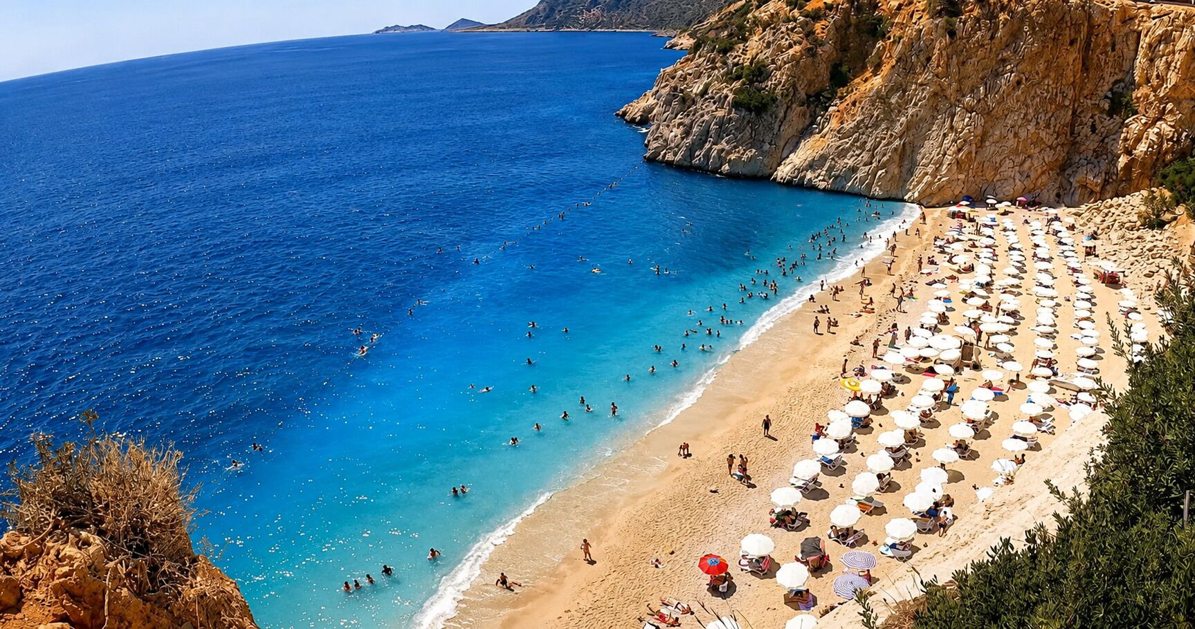 Una spiaggia con numerosi lettini e ombrelloni, una costa rocciosa e acque blu cristalline.
