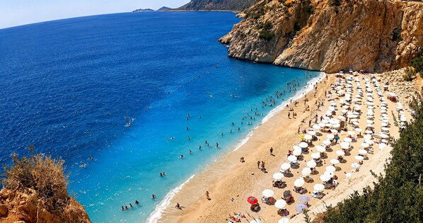 A beach with numerous sunbeds and umbrellas, a rocky coastline, and clear blue water.