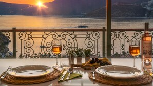 Une table dressée pour deux personnes, avec des verres à vin, des assiettes et une bouteille de vin rosé, donnant sur une étendue d’eau au coucher du soleil.