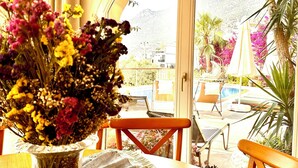 Un coin repas avec une table dressée pour deux personnes, un vase de fleurs et une vue sur une piscine et des montagnes, visible à travers une porte vitrée.
