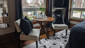 A hotel room with two chairs, a small table, a bed, a TV, and a window with a view of buildings and trees. - Stonewater Manor (Gananoque)