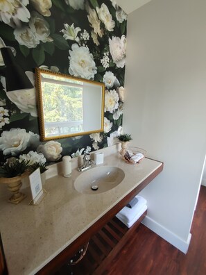 A bathroom with a floral wallpaper, a gold-framed mirror, a sink, and a small potted plant. - Stonewater Manor (Gananoque)