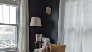 A room with a dark wall, a window with white curtains, a lamp, a side table with a pitcher, and a sofa with a throw blanket. - Stonewater Manor (Gananoque)