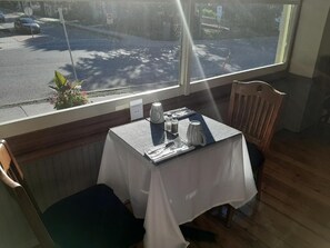 A dining area with a table set for two, a window view of a street, and a plant on the windowsill. - Stonewater Manor (Gananoque)