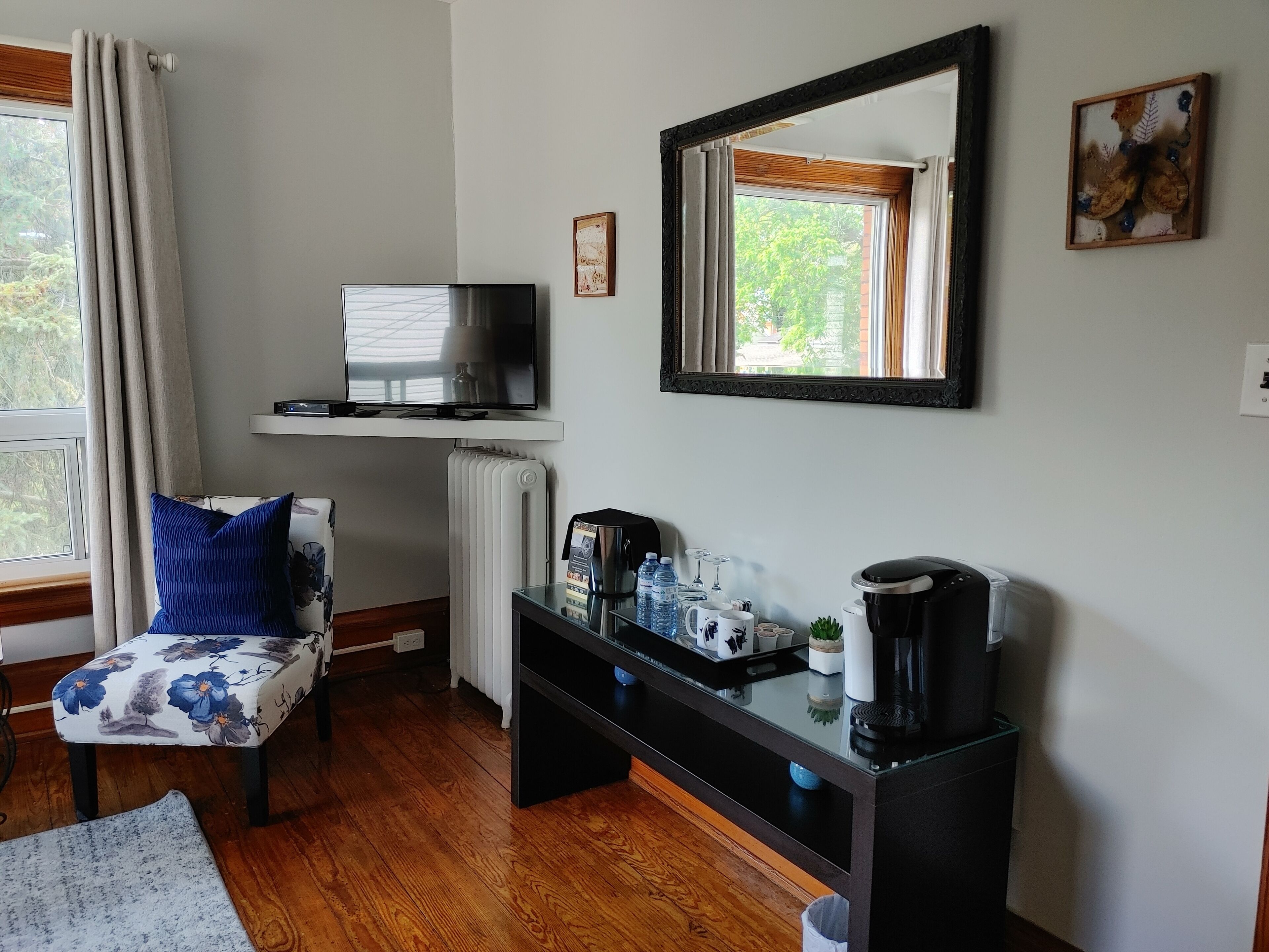 Una habitación con suelo de madera, una silla con cojín estampado, una estación de café con tetera y tazas, un televisor sobre una estantería y una ventana con cortinas.