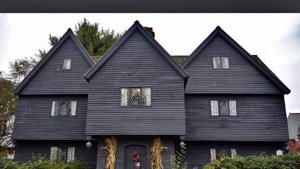 Et mørkt, toetages hus med symmetrisk design, der har en midterdør omkranset af to vinduer, og en lille veranda med dekorative detaljer.