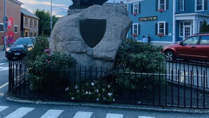 En statue af en figur i kåbe på en stenfundament med et skjold, omgivet af et hegn og blomster, foran en bygning med skilt, der siger "Stepping Stone Inn".