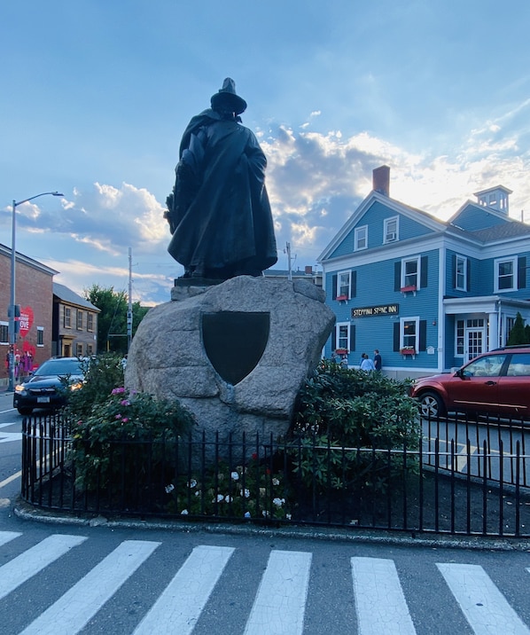 Una estatua de una figura con capa sobre un pedestal de piedra con un escudo, rodeada por una valla y flores, frente a un edificio con un letrero que dice "Stepping Stone Inn".