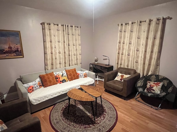 A living room with a sofa, armchair, coffee table, and a rug. There is a desk with a lamp and a chair.