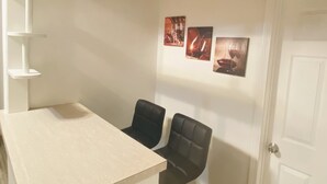 Et lille, kompakt værelse med et hvidt skrivebord og to sorte stole. Tre billeder hænger på væggen, og en hvid dør er synlig.