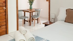 Quarto com cama, porta de madeira, mesa de jantar com cadeiras e uma mesinha com abajur.