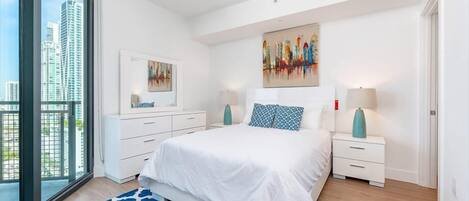 A modern bedroom with a large bed, a city view, a painting on the wall, and a patterned rug.