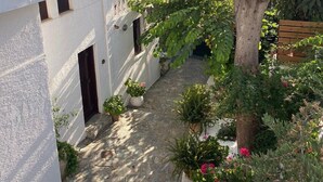 A narrow, white-walled courtyard with a stone pathway leading to a door. There are potted plants and a tree with green foliage. - Troullos Traditional Houses (Archanes-Asterousia)