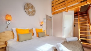 Une chambre avec une tête de lit en bois, un lit avec une literie blanche et orange, une lampe fixée au mur et une échelle en bois menant à une mezzanine.
