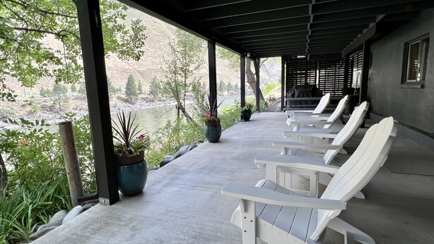A covered outdoor seating area with white Adirondack chairs, potted plants, and a view of a river and hills. - The Building on The River R&R  (Riggins)