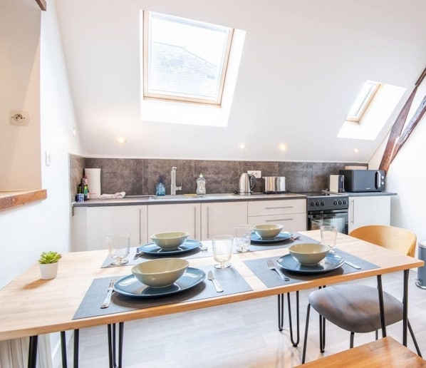 Une cuisine moderne avec un coin repas, comprenant une longue table dressée pour quatre personnes, un four encastré et une grande fenêtre au-dessus de l’évier.