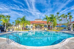 Una piscina con cielo azul despejado, rodeada de palmeras y un edificio con techo rojo.