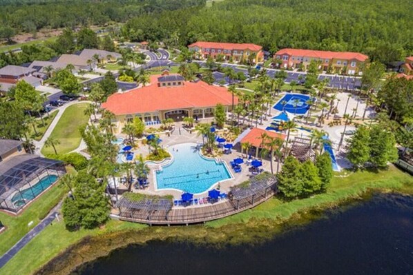 An aerial view of a resort with a central swimming pool, multiple buildings, and a surrounding natural landscape. - Magical 7BR Resort Pool Home Near Disney World (Kissimmee)