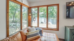 A room with a large window overlooking a natural landscape, a leather sofa with patterned cushions, and a wall-mounted light fixture.