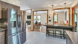 A modern kitchen with a stainless steel refrigerator, a dining area with a wooden table and chairs, and a large window providing natural light.