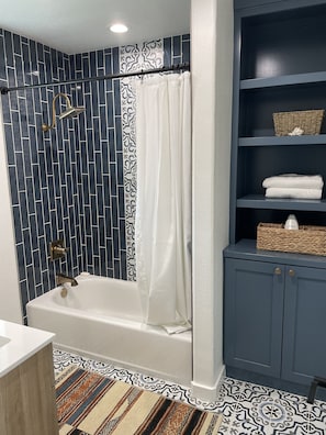 A modern bathroom with a bathtub, a shower with a gold faucet, and a blue tiled wall. There are shelves with towels and baskets.