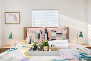 Ein ordentlich bezogenes Bett mit farbenfroher Decke, zwei Nachttischlampen, eine gerahmte Karte an der Wand und ein Fenster mit Jalousien.