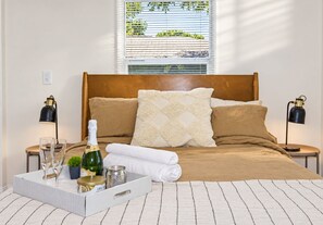 A neatly made bed with a wooden headboard, two bedside lamps, a window with blinds, and a tray with a bottle, glasses, and a candle. - Perfect & Cozy Home in A+ Location (Saint Petersburg)