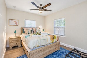 A bedroom with a wooden bed, a nightstand, a desk lamp, a ceiling fan, and two windows with blinds.