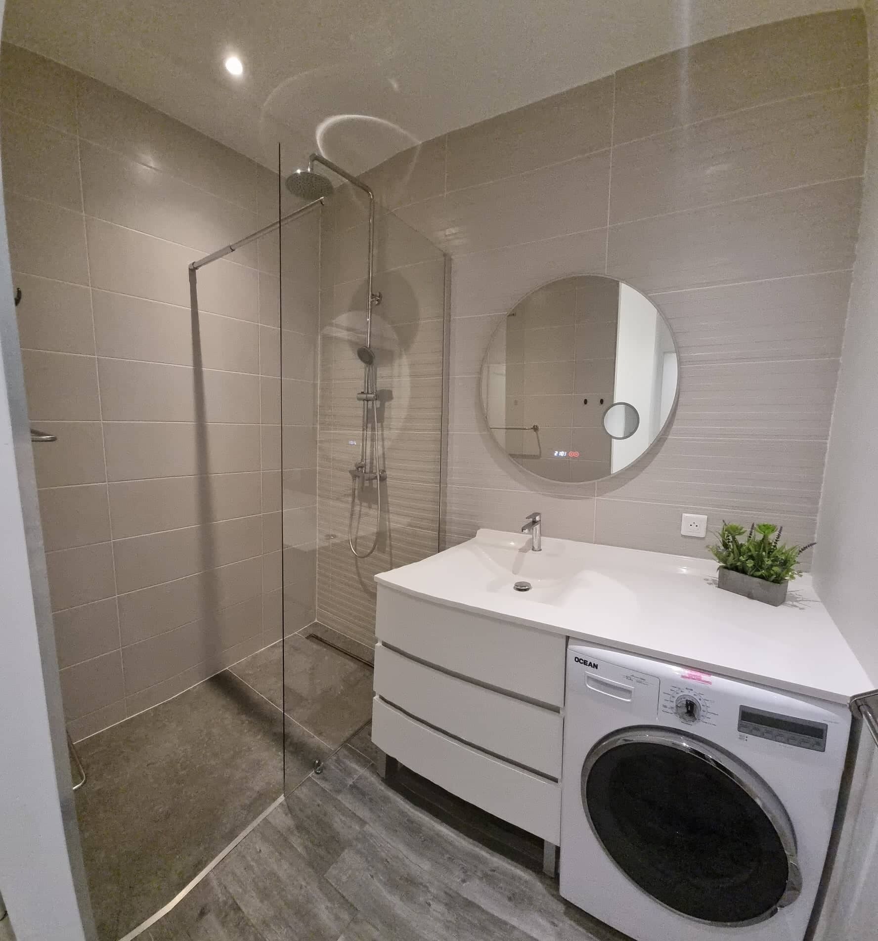 Une salle de bain moderne avec une douche à parois de verre, un miroir rond, un lavabo blanc agrémenté d’une petite plante et un lave-linge à chargement frontal.