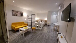Une chambre d’hôtel compacte avec un canapé jaune, une petite table blanche, un lit avec une tête de lit en bois, un téléviseur fixé au mur et une porte coulissante.