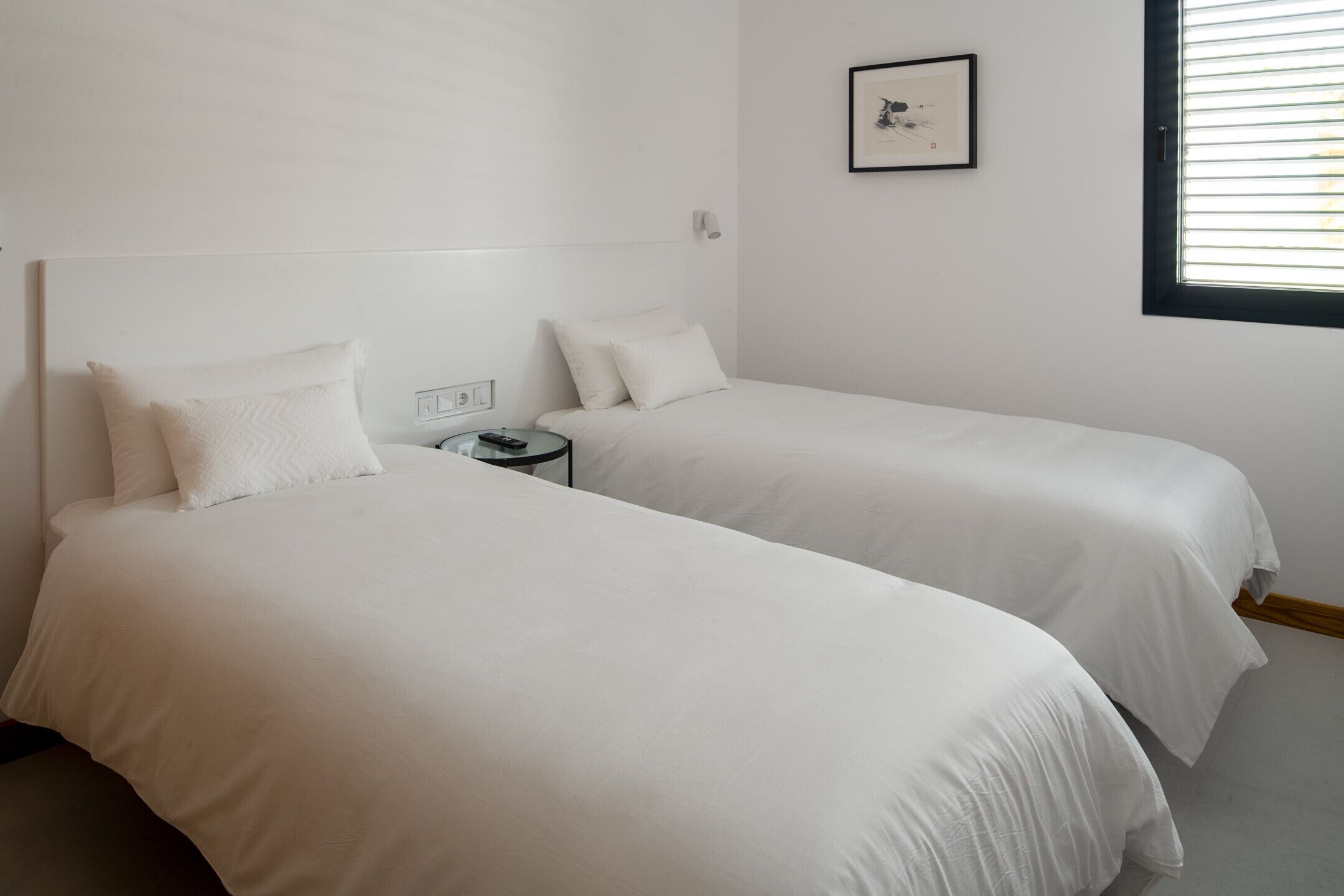 A twin room with two beds, a small bedside table, a framed picture on the wall, and a window with blinds.