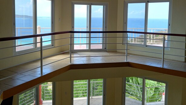 Un intérieur à plusieurs niveaux, doté de grandes fenêtres offrant une vue sur l’océan, d’un ventilateur de plafond et d’un balcon avec une balustrade.