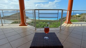 Un espace extérieur abrité, doté d’une table et d’un verre, offrant une vue panoramique sur la mer et les collines.