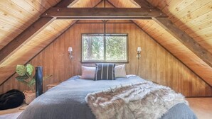 Une chambre mansardée chaleureuse, avec un plafond en bois incliné, une grande fenêtre et un lit agrémenté d’un oreiller moelleux.