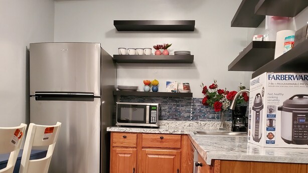 A compact kitchen with a stainless steel refrigerator, a microwave, wooden cabinets, and a small island with a sink. - Cosy Studio in a Private home with private entrance and door (Bronx)