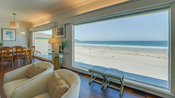 A room with a large window overlooking a beach, a dining area with wooden furniture, and a seating area with a bench. - Blue Sea Villa, Centered in La Jolla Shores! (San Diego)