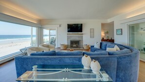 A living room with a large blue sofa, a fireplace, a flat-screen TV, and a view of the beach. - Blue Sea Villa, Centered in La Jolla Shores! (San Diego)
