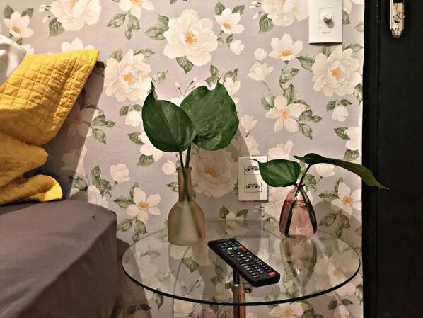 Une table en verre, deux plantes en pot, une télécommande et une couverture jaune sur un lit.