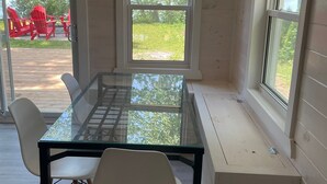 A room with a glass-top table, white chairs, and large windows offering views of outdoor seating and greenery. - Modern Cottage on the Water - Newly Renovated! (Dashwood)