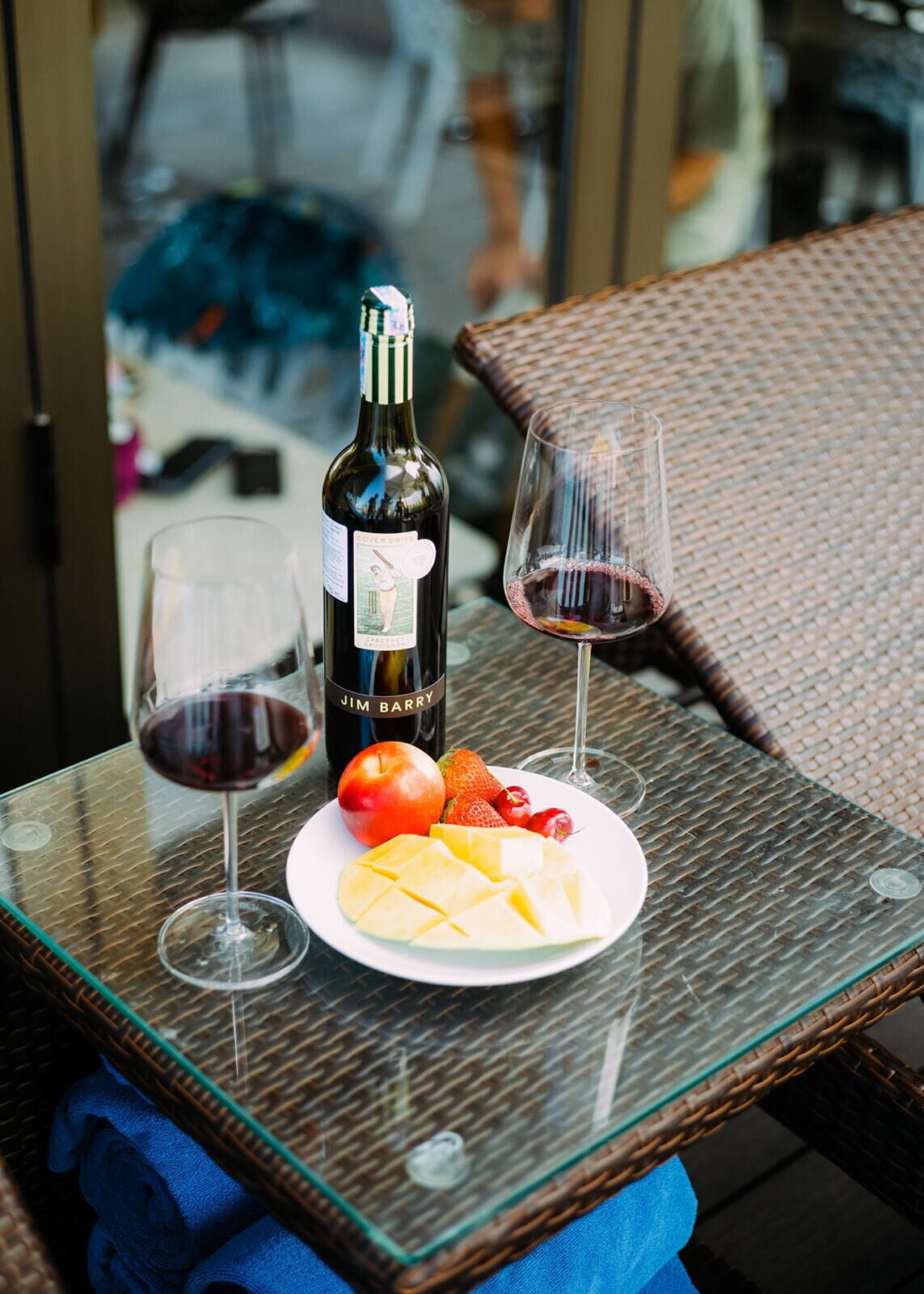 Une table en verre avec une bouteille de vin, deux verres à vin et une assiette de fruits.