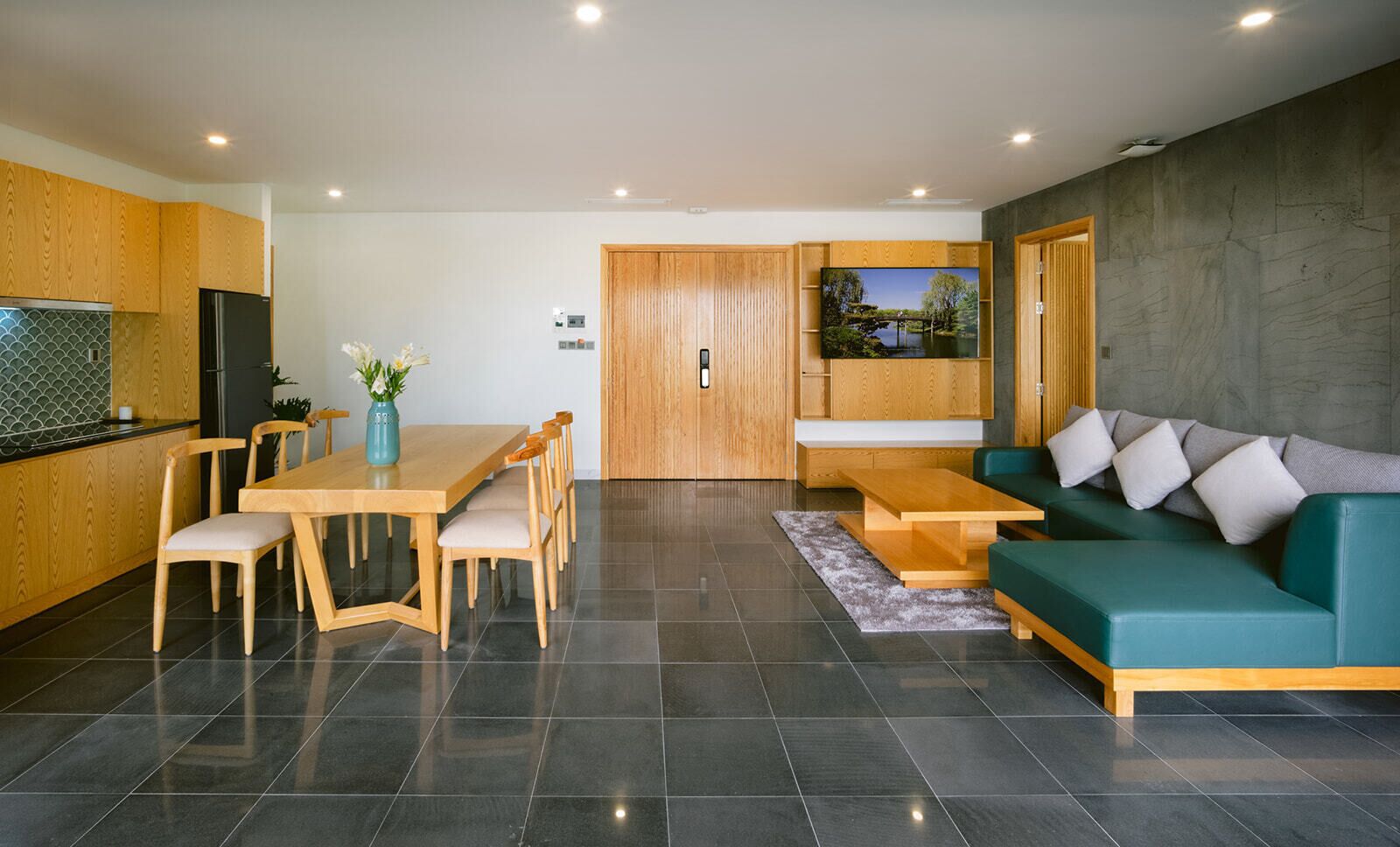 Een moderne keuken met houten kastjes, een zwarte koelkast en een eettafel met stoelen. Er staat een groene bank en een televisie hangt aan de muur.