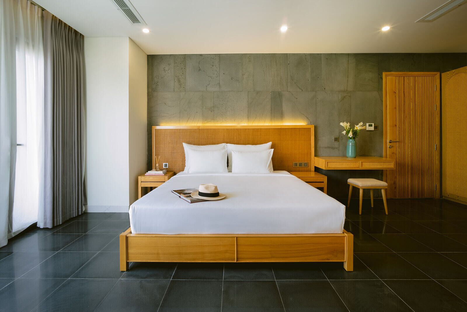 A modern hotel room with a large bed, wooden headboard, a nightstand, a desk with a chair, and a wardrobe.