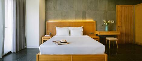 A modern hotel room with a large bed, wooden headboard, a nightstand, a desk with a chair, and a wardrobe.