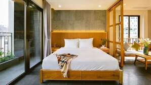 A modern hotel room with a large bed, a wooden headboard, a bedside table, a nightstand, a window with a view of buildings, and a balcony with plants. - LIVIE DANANG ANTHUONG (Da Nang)