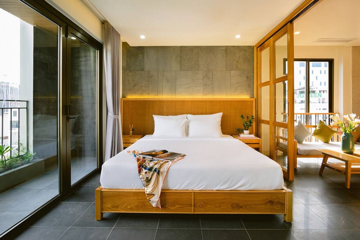 Ein modernes Hotelzimmer mit einem großen Bett, einem Holzkopfende, einem Nachttisch, einem Fenster mit Blick auf Gebäude und einem Balkon mit Pflanzen.