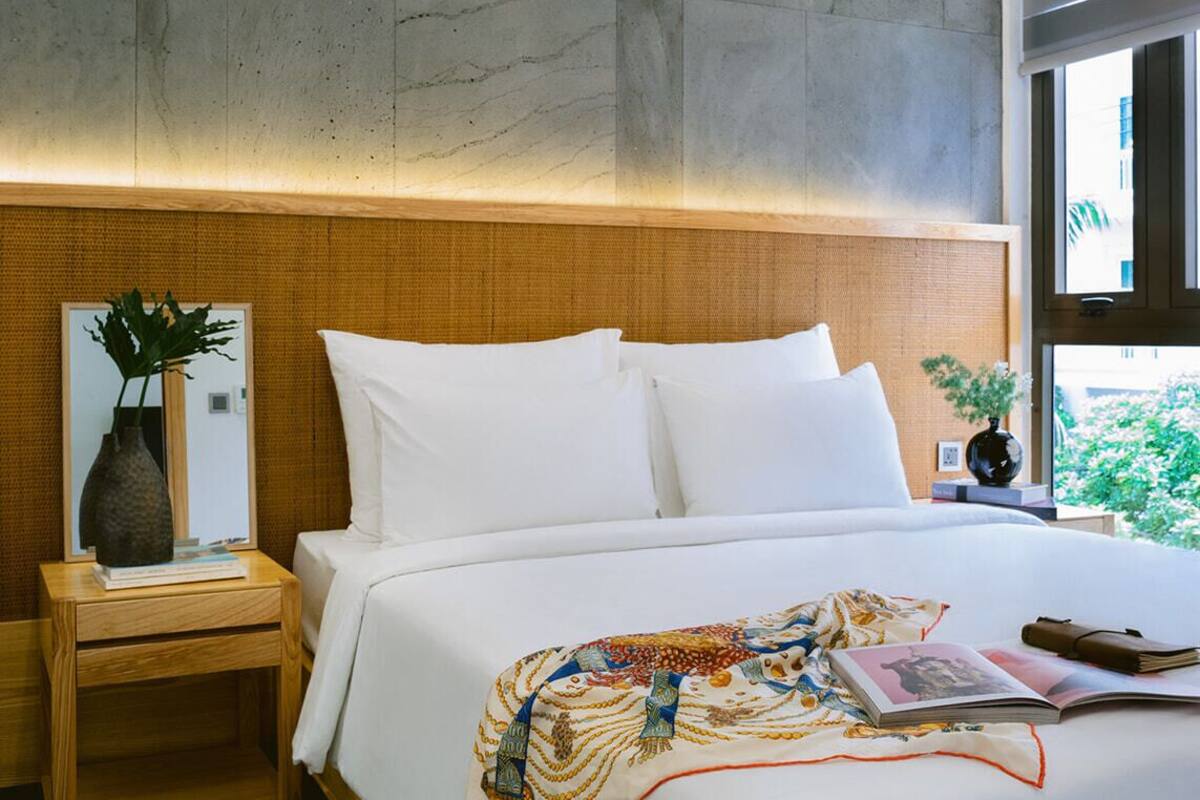 Ein modernes Hotelzimmer mit einem großen Bett, einem Holz-Kopfstück, einem Nachttisch mit Vase und Buch sowie Blick auf Grünflächen.