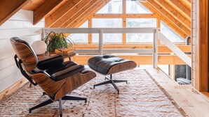 A cozy loft bedroom with a sloped ceiling, a large window, a wooden ceiling, a black leather chair, a matching ottoman, and a small table with a plant and a book.