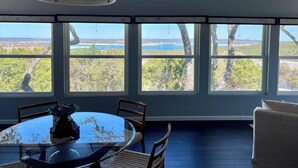 Ein Essbereich mit einem runden Glastisch, Stühlen und einer Pendelleuchte. Große Fenster bieten Blick auf Bäume und einen entfernten Strand.