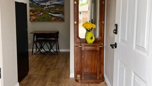 A hallway with wooden flooring, a bench, a painting on the wall, and a door with a mirror and a vase with flowers. - Cozy, modern suite in the heart of Loveland Colorado. (Loveland)